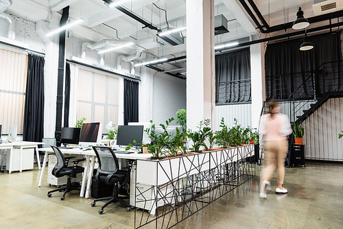 Motion blur of businesswoman walking near computers and plants in office