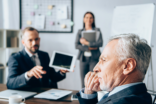 Pensive investor sitting near business people on blurred background in office