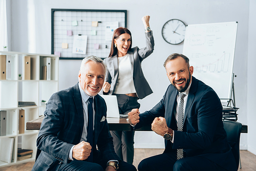 Cheerful businessmen showing yeah gesture near colleague on blurred background in office
