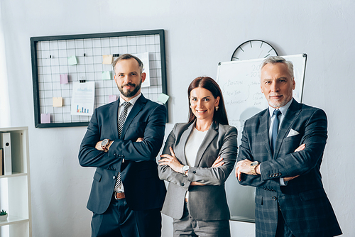 Smiling businesswoman standing with crossed arms near partners in office
