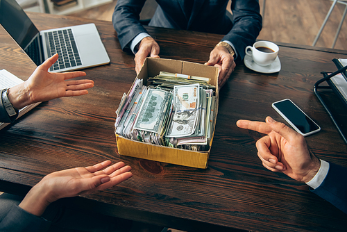 Cropped view of businesspeople pointing at box with money near colleague and devices on blurred foreground