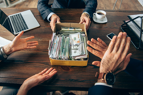 Cropped view of businessman holding box with cash near colleagues showing stop gesture on blurred foreground