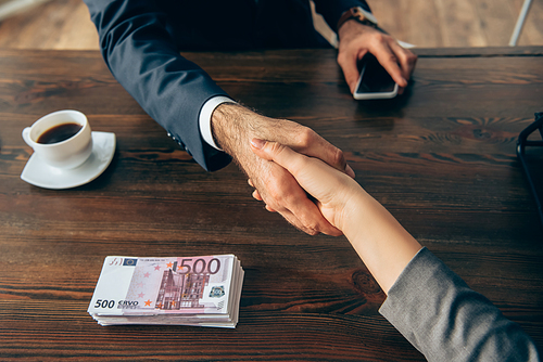 Cropped view of business partners shaking hands near coffee and euro banknotes on table