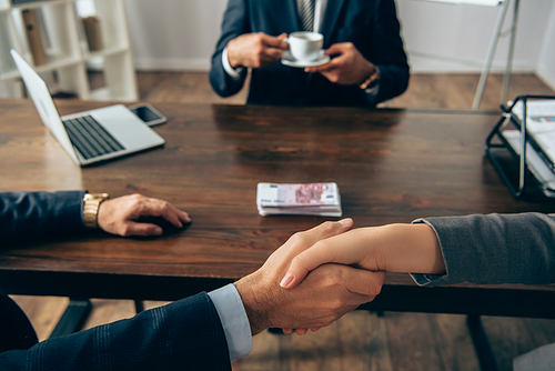 Cropped view of business partners shaking hands near money and colleague  with cup on blurred background