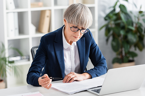 middle aged team leader in glasses holding pen near documents on desk