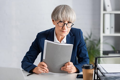 middle aged team leader in glasses holding folder near gadgets and paper cup on desk
