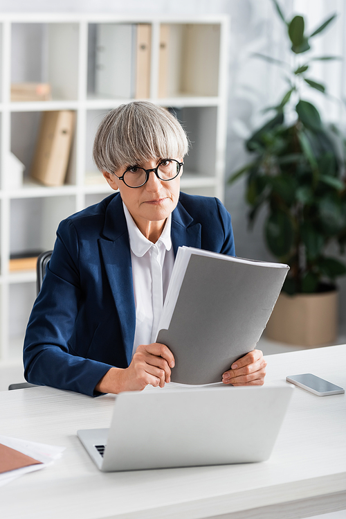 middle aged team leader in glasses holding folder near gadgets on table