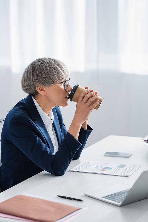 middle aged team leader in glasses drinking coffee to go near papers with charts and graphs on desk