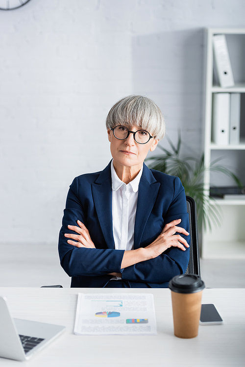 middle aged businesswoman in glasses sitting with crossed arms near charts and graphs on desk