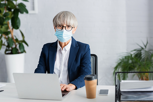middle aged team leader in glasses and medical mask using laptop near smartphone and paper cup on desk
