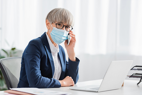 middle aged team leader in glasses and medical mask talking on mobile phone near laptop on desk