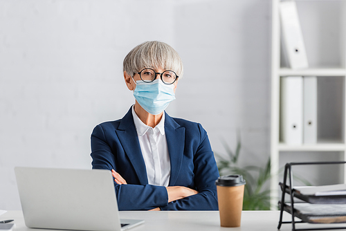 middle aged team leader in glasses and medical mask sitting with crossed arms near laptop and paper cup on desk