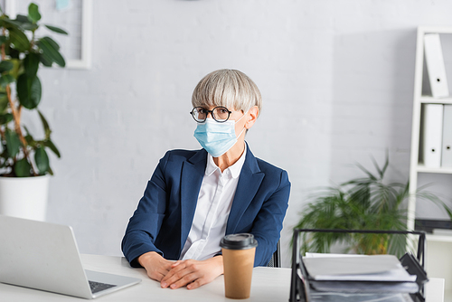mature team leader in glasses and medical mask  near laptop and paper cup on desk
