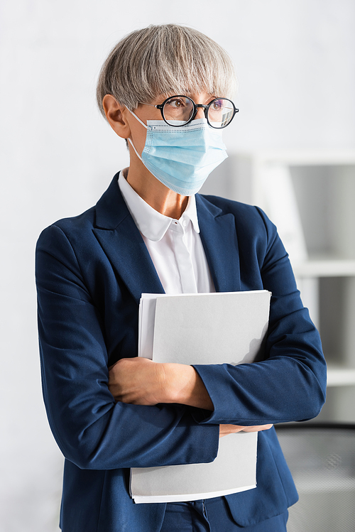 mature team leader in glasses and medical mask holding folder in office