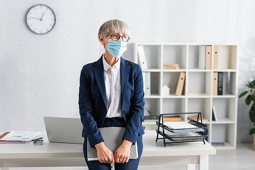mature team leader in glasses and medical mask holding folder while standing near workplace