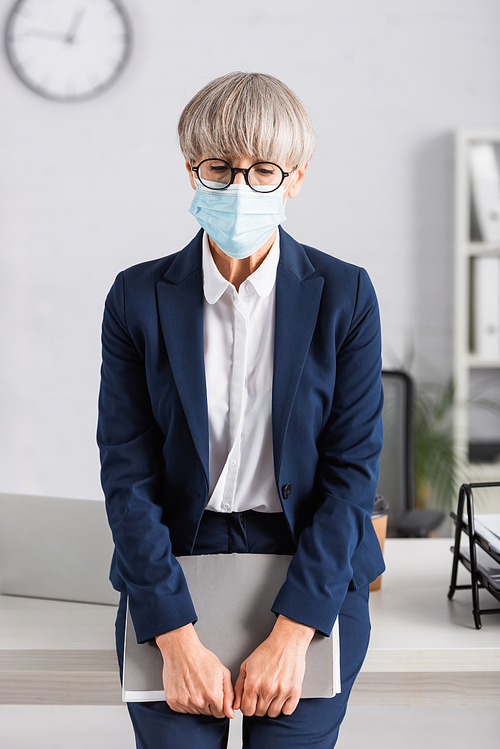 middle aged team leader in glasses and medical mask holding folder while standing near workplace