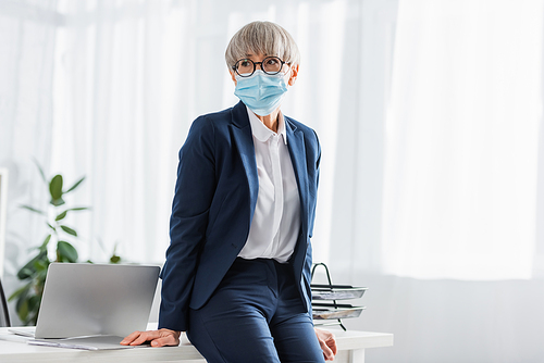mature team leader in glasses and medical mask leaning on desk with laptop in office