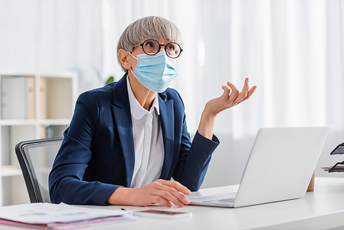 mature team leader in glasses and medical mask gesturing while looking up in office