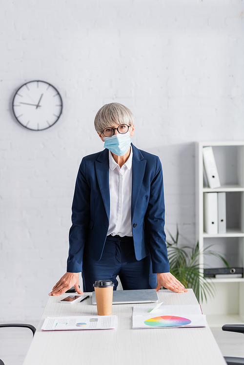 middle aged team leader in glasses and medical mask standing near workplace in modern office