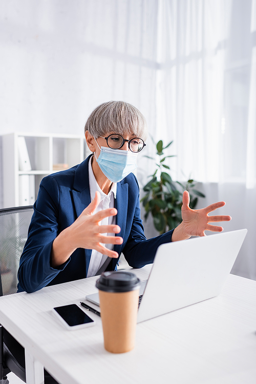 mature team leader in glasses and medical mask gesturing during video call in office
