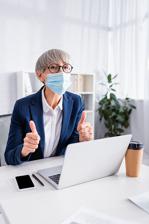 mature team leader in glasses and medical mask showing thumbs up near gadgets on desk