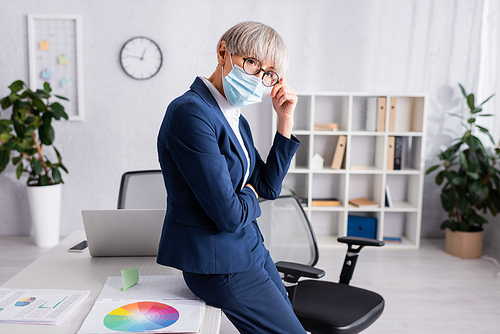 mature team leader in glasses and medical mask leaning on chair near desk