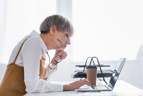 side view of mature team leader holding glasses and using laptop in office