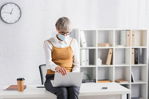 middle aged team leader in glasses and medical mask using laptop while sitting on desk