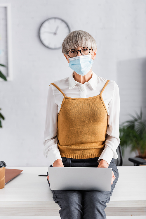 middle aged team leader in glasses and medical mask using laptop while sitting on desk in office