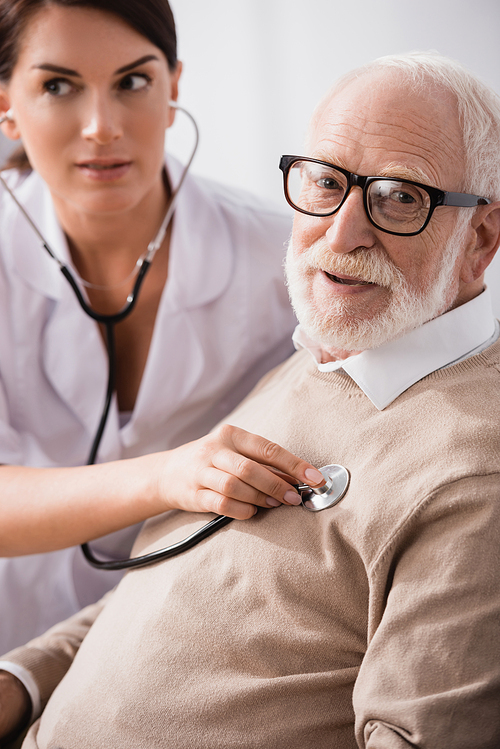 medical assistant examining aged man with stethoscope on blurred background