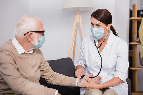 social worker in medical mask examining elderly man with stethoscope at home