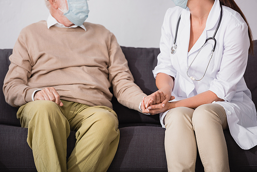 partial view of social worker in medical mask holding hands of aged man at home