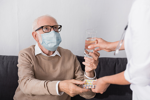 elderly man in medical mask taking glass of water and pills from geriatric nurse on blurred foreground