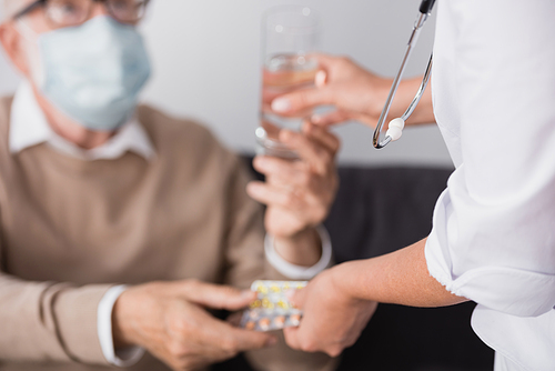 cropped view of social worker giving medicines and water to elderly man on blurred background