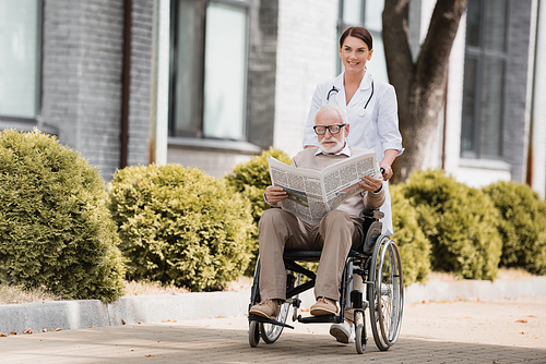 cheerful social worker walking with disabled elderly man reading newspaper in .