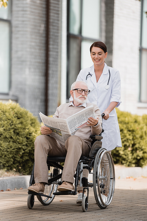 smiling geriatric nurse walking with aged disable man holding newspaper in . outdoors