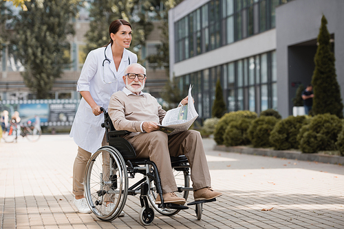 geriatric nurse walking with elderly disabled man in . with newspaper outdoors