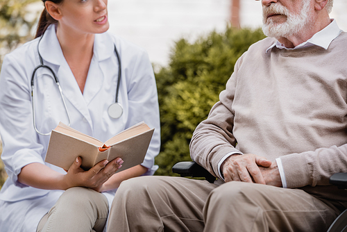 cropped view of geriatric nurse reading book to elderly man outside