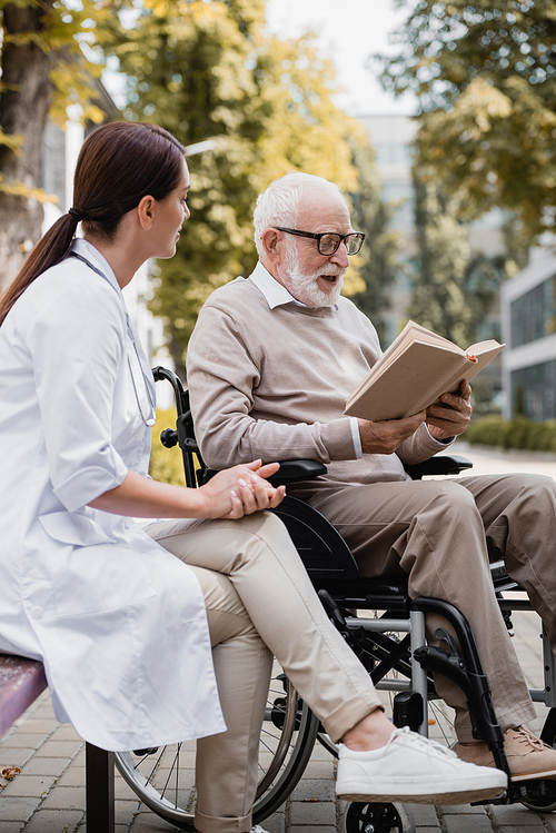 geriatric nurse sitting near aged disabled man reading book outdoors