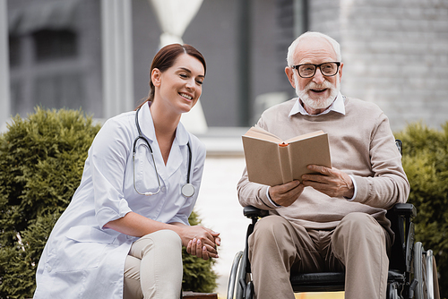 cheerful geriatric nurse near aged disabled man reading book in . outdoors