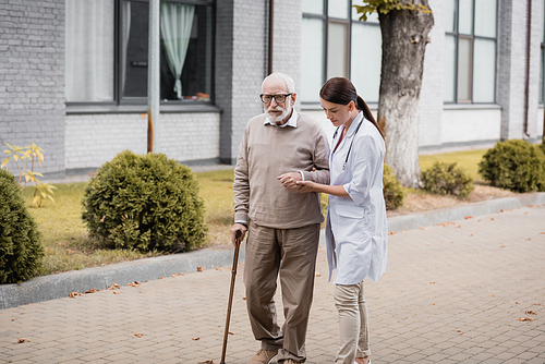 social worker supporting man with walking stick while strolling together outside