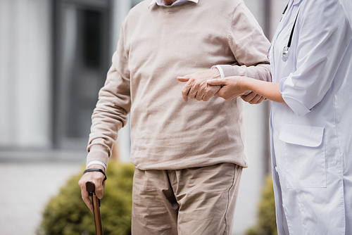 cropped view of social worker supporting aged man strolling with walking stick