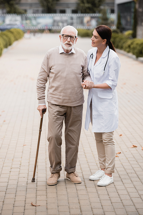 social worker supporting aged man with walking stick while strolling together
