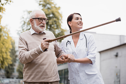 aged man pointing with walking stick away near smiling geriatric nurse