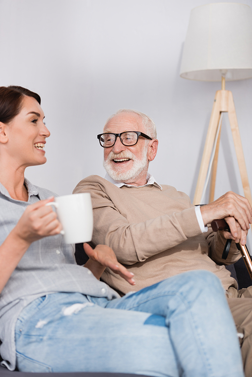 smiling woman holding tea while talking to aged father on blurred foreground