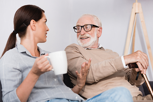 brunette woman holding tea and gesturing while talking to cheerful elderly father