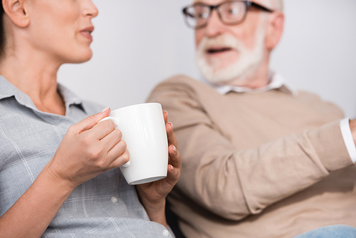 woman holding cup of tea while talking to surprised father on blurred background