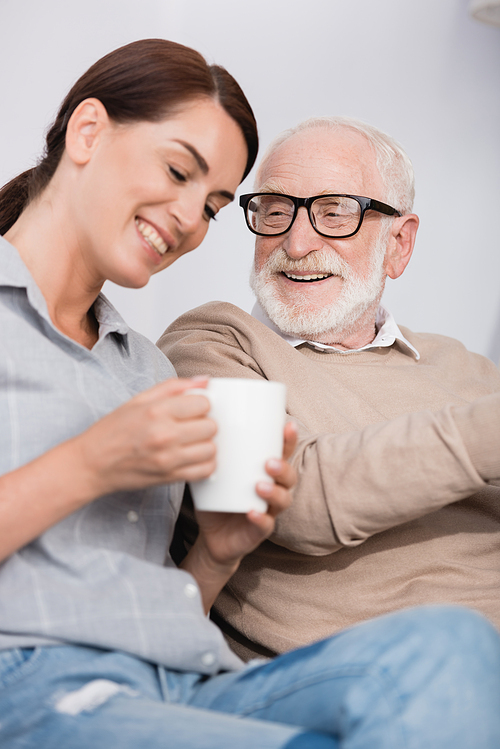 cheerful woman with cup of tea talking to aged smiling father on blurred foreground