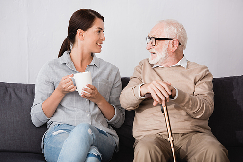 brunette woman with cup of tea and smiling aged man with walking stick talking while sitting on sofa at home