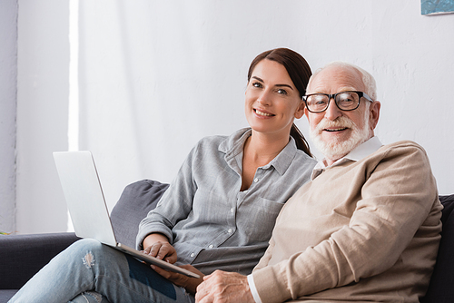 smiling brunette woman  while sitting near aged father and using laptop